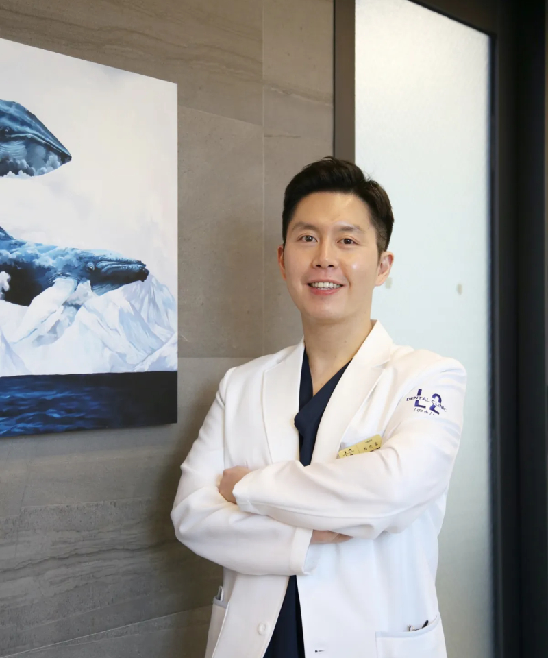 엘투치과 최순호 대표원장 프로필 이미지 흰색 진료복을 입은 치과의사가 고래 그림 앞에서 팔짱을 끼고 미소 짓고 있는 모습